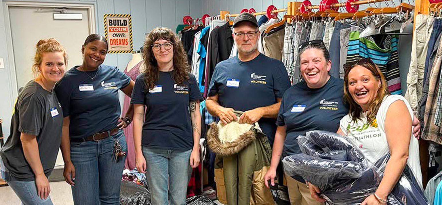 Six smiling adults wearing volunteer shirts and name tags stand together in a room of hanging clothes, posing by racks of donations. A motivational sign hangs behind them, inspiring others to donate and volunteer to support youth in need.