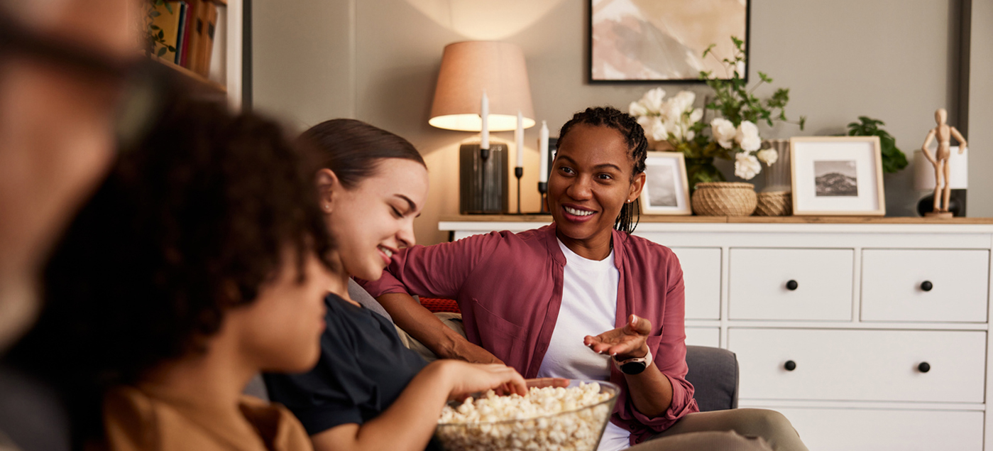 A group of people sit on a sofa in a cozy living room, smiling and talking while sharing a large bowl of popcorn. The background features framed pictures, flowers, and a lamp on a white dresser.